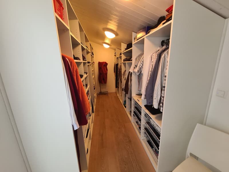 A narrow closet with wooden floors, white shelves on both sides, and several clothing items organized in the racks and baskets. Ceiling is white with recessed lights. A metal stand with a red cloth is present.
