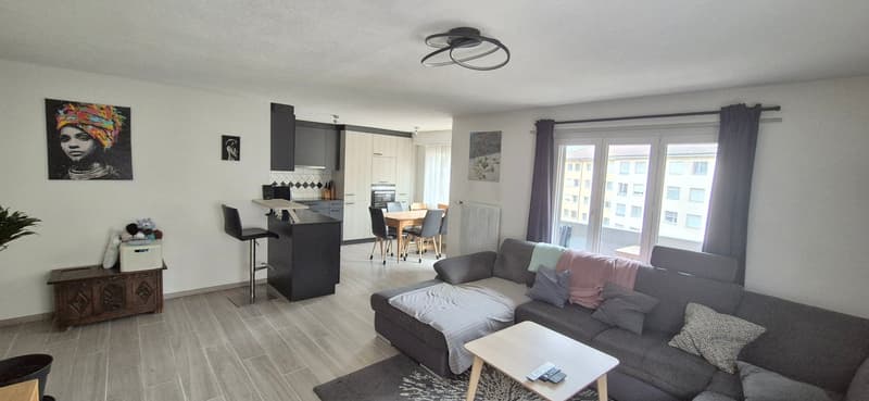 Open plan living and dining area with wooden flooring, grey couch, coffee table, bar counter, kitchen island, breakfast table, radiator, dining chairs, side table, carpet, decorative painting, and window curtains.