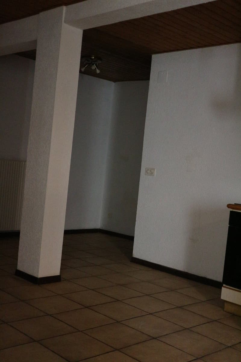 The image shows a dark, empty room with white walls and a tiled floor. There is a light fixture visible on the ceiling, and the room appears to be a basement or storage area.