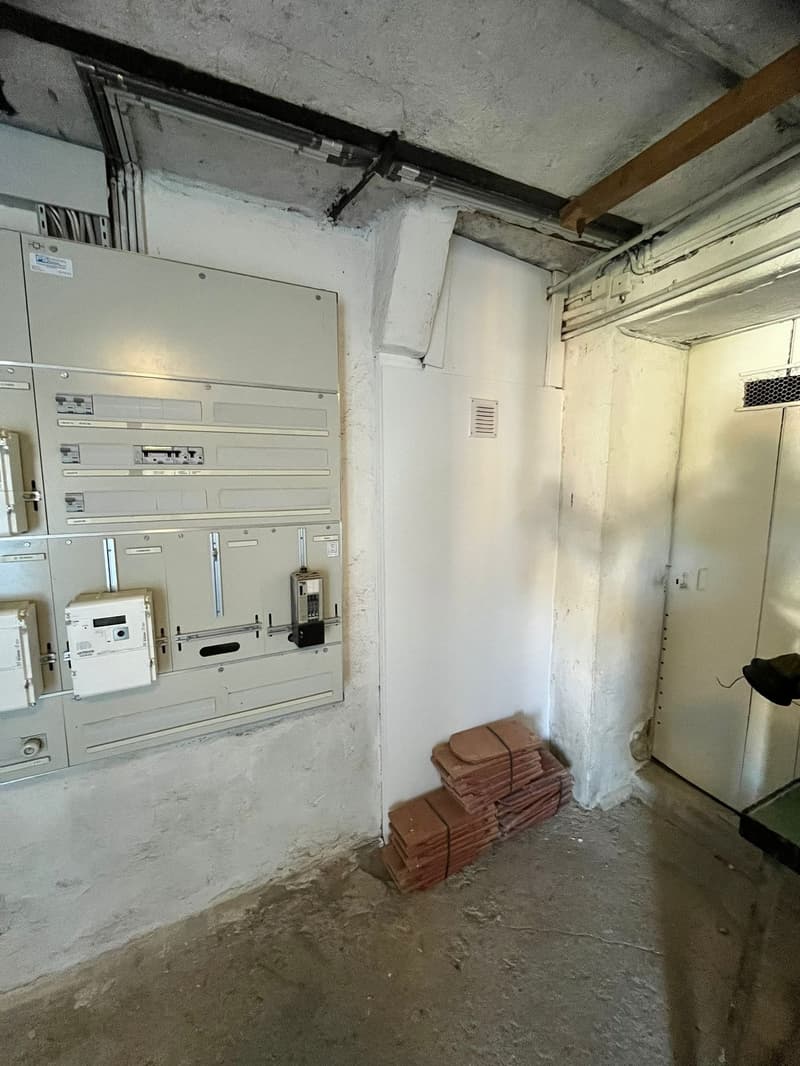The image shows a utility room or storage area with concrete walls and floor. There is a large electrical panel on the wall, and a stack of tiles or other building materials on the floor.