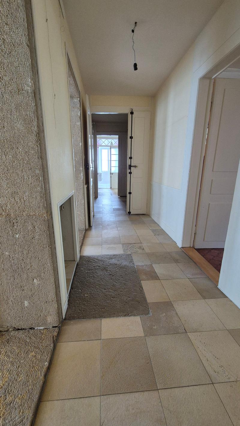An empty hallway with tiled flooring, white walls, and wooden doors. A carpet on the floor, a window at the end, and a lamp hanging from the ceiling. Two rooms with wooden doors and one room with a white door.