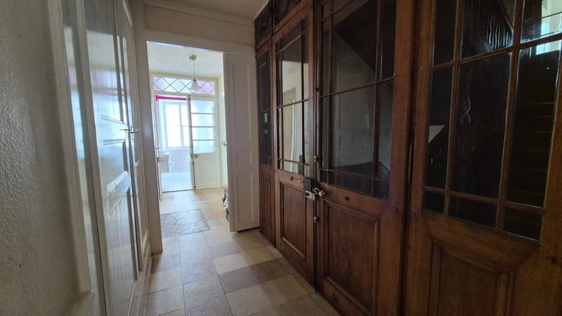Corridor with wooden doors, checkered tiles, white walls, open doorway leading to a bathroom. A small rug on the floor. A staircase with steps visible behind the wooden doors.