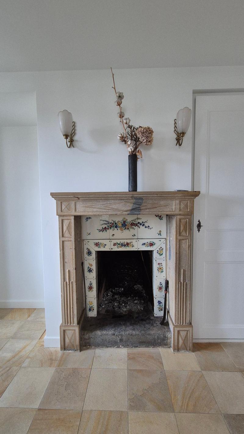 Fireplace with wooden frame, tiled hearth, and decorative tiles; above fireplace is a mantel with flowers, two wall-mounted lamps on each side, and a white wooden door.