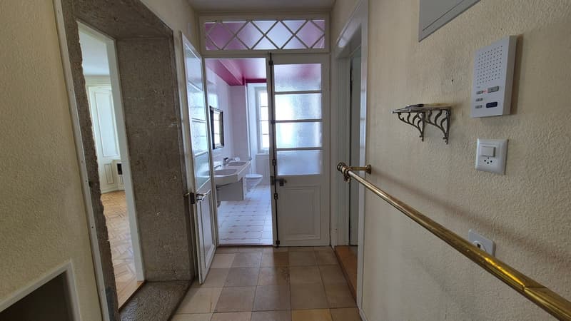 White walls, tiled floor, open doorway leading to bathroom, dual sinks, and toilet visible through door, bathroom has pink ceiling, mirror, and tiled floor, decorative window.