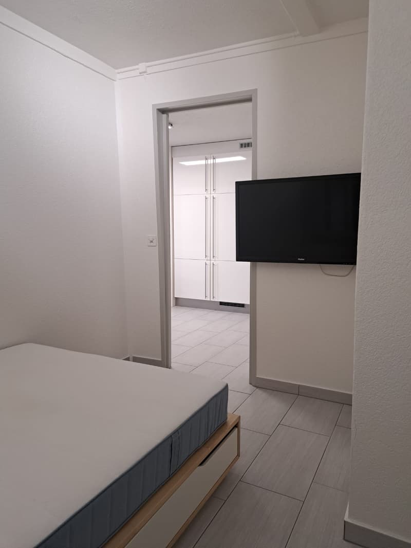 White wall, wooden bed, tiled floor, TV on the wall, open door