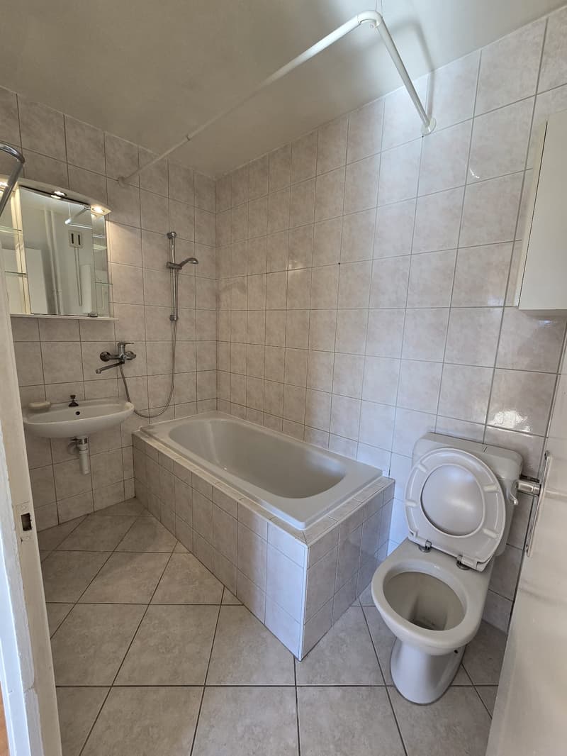 A clean and well-lit bathroom with a bathtub, a toilet, a sink, and a mirror. It has white walls and tile flooring.