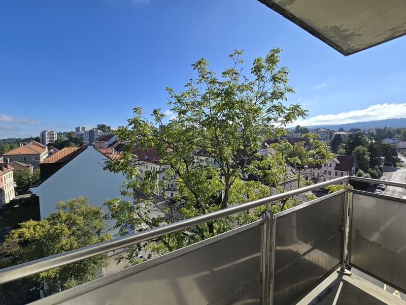 Balcony with metal railings, large tree in the middle, cityscape in the background, blue sky, distant hills