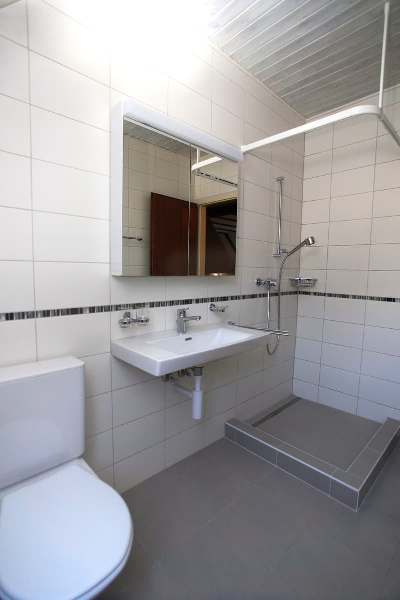Modern bathroom, tiled floor, glass door shower, toilet, washbasin, and mirror with lighting