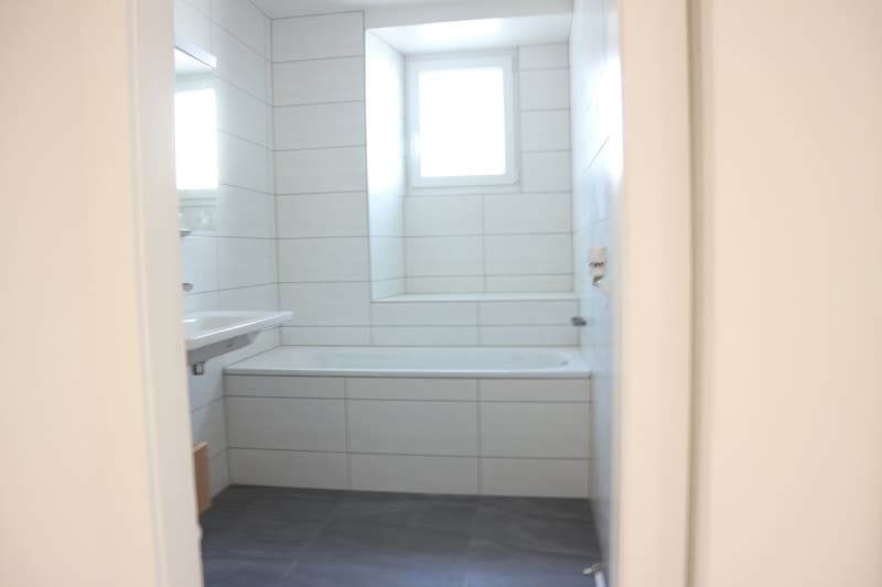 A modern, minimalist bathroom with white tiles on the walls and floor, a bathtub, a sink, and a window
