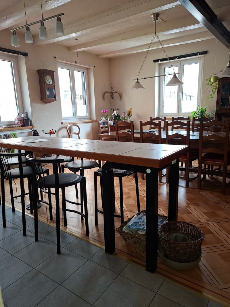 long table with chairs, 4 chandeliers, wooden floor, white walls, 2 windows, vintage clock on wall, side tables, plants