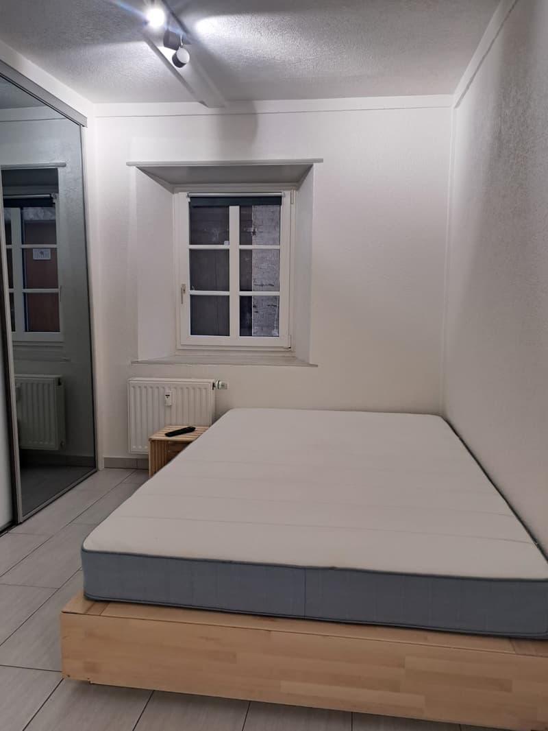 Spacious bedroom with a double bed, white walls, ceiling lamps, a small nightstand, a window with a view, and two radiators.