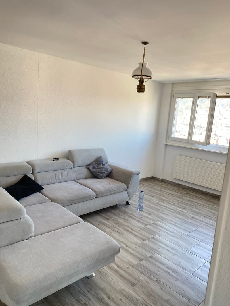 L-shaped sofa, wooden floor, white walls, large window, radiator, bottle on the floor