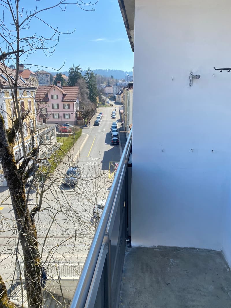 Balcony with metal railing, overlooking a street with cars parked, buildings, and a tree.