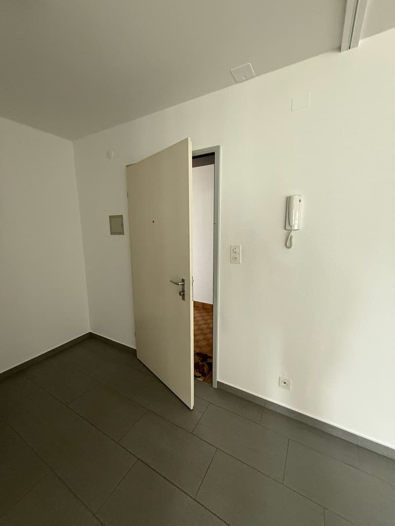 empty room, white walls, gray tile flooring, open door, telephone on the wall, power outlet