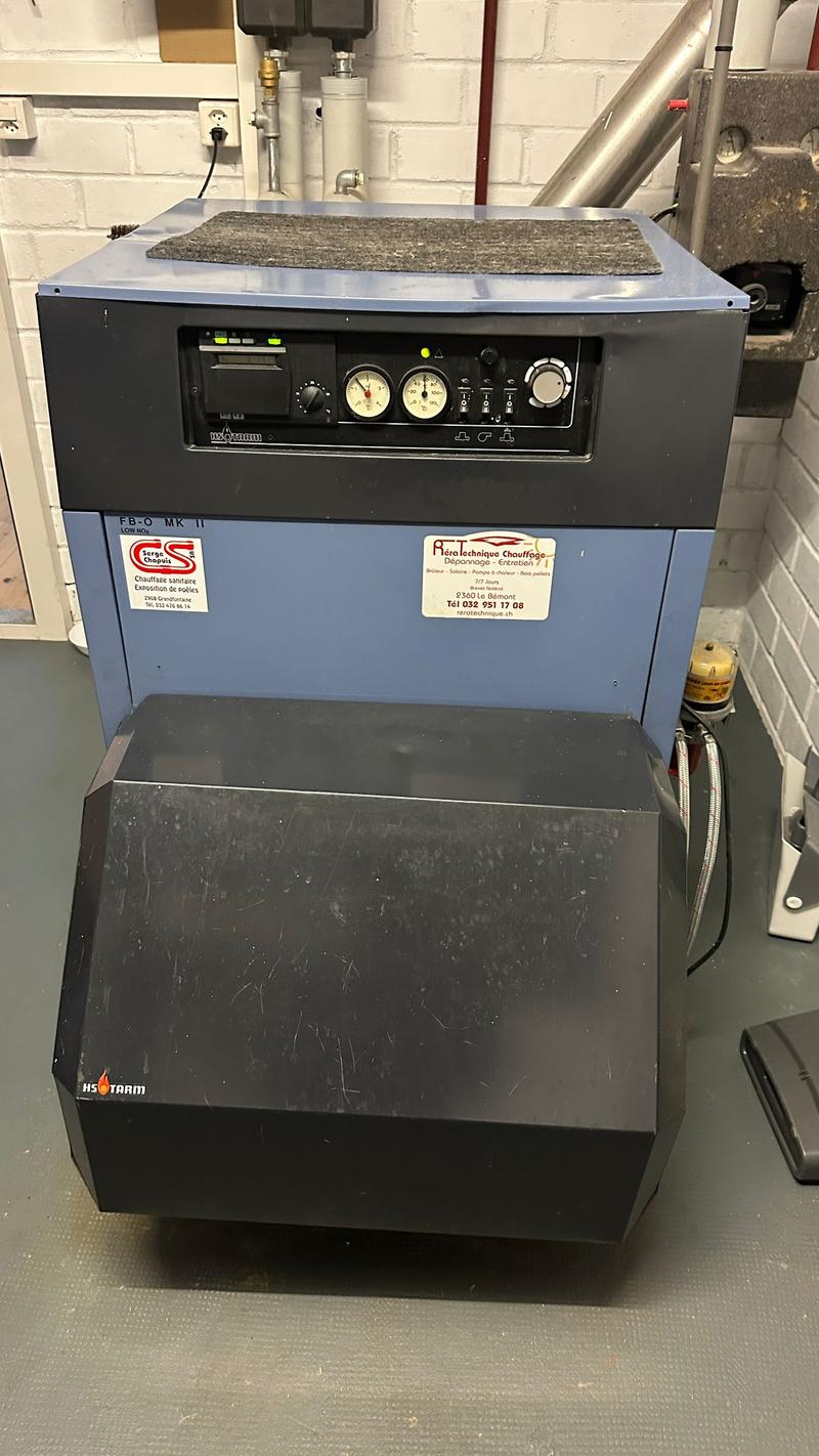 An industrial machine with a large gray mat on top, a black front, and a blue side. The machine has a series of dials, buttons, and gauges on the front, and a large black rectangular compartment at the bottom. The machine is placed on a tiled floor.