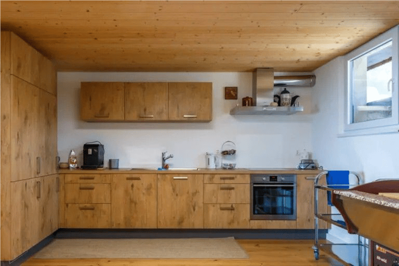Modern open kitchen with wooden cabinets, sink with faucet, oven, coffee maker, kettle, towels, and other appliances. Wooden floor, white walls, ceiling, a window.