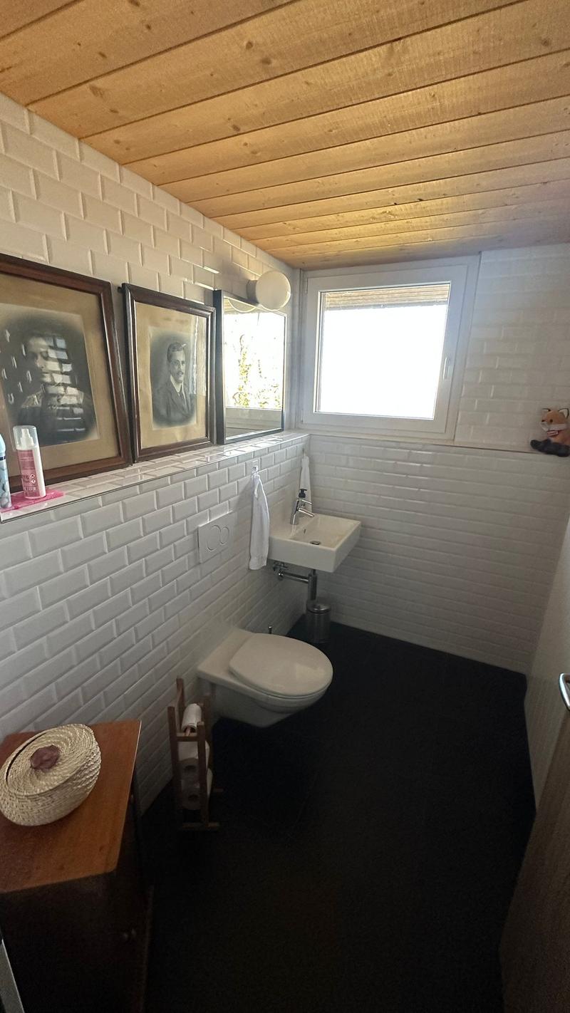 small compact bathroom with white walls, white sink, white toilet, mounted picture frames, toilet paper holder, wooden side table with decorative object