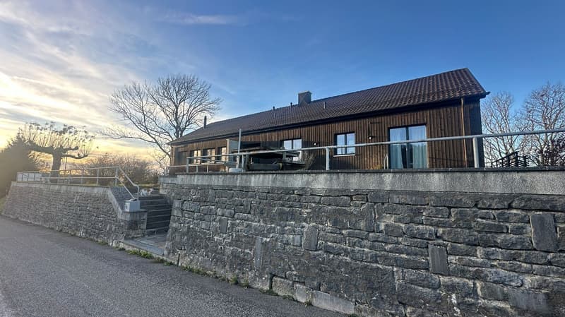 2 story wooden house, elevated ground, brown roof, large windows, staircase to front, stone wall