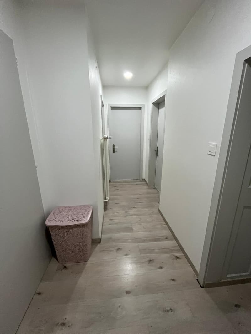 corridor with wooden flooring and white walls, a trash bin is present