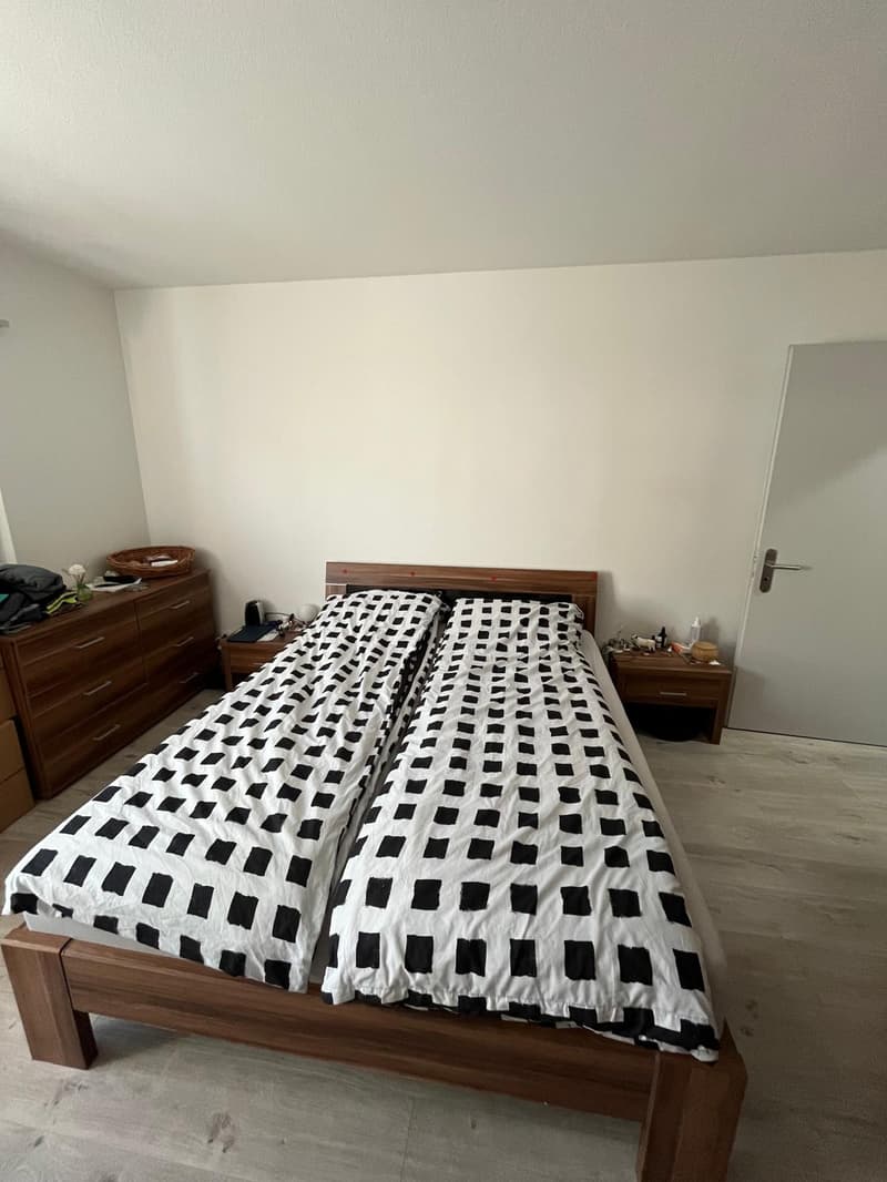 Bedroom with two single beds, wooden bedframe, white and black bed sheets, wooden nightstands with lamps, wooden dresser with drawers, white walls.