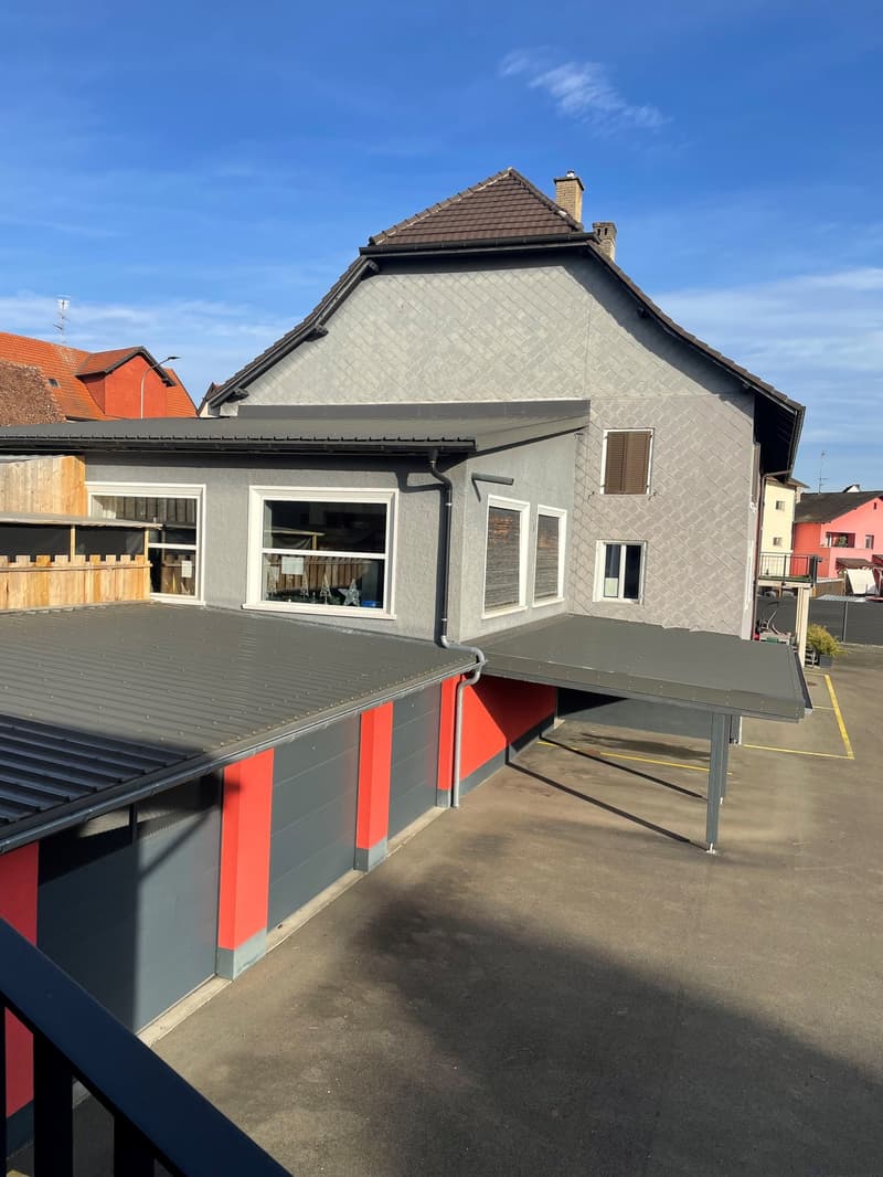 two story house, grey exterior with red roof, several windows, covered parking area, fence, adjacent wooden house