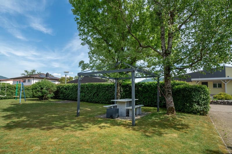 Grassy lawn with a large tree, a covered gazebo structure with a table and benches, and a playground equipment in the background.