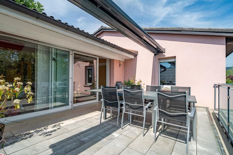 Two-story house with pink exterior walls, large windows, and a covered patio area with a dining table and chairs. The patio has a tiled floor and overlooks a garden or outdoor space.
