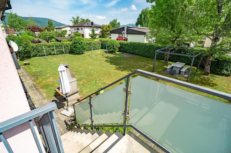 2-story residential building with a balcony, surrounded by a well-maintained garden with a playground and outdoor furniture. The building is situated in a lush, green environment with trees and mountains in the background.