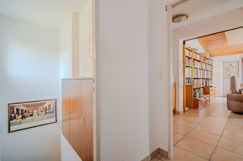 The image shows the interior of an apartment. The space includes a hallway with a framed artwork on the wall, leading to a living room area with bookshelves and a sofa. The flooring appears to be tile, and the walls are painted a light color. The overall