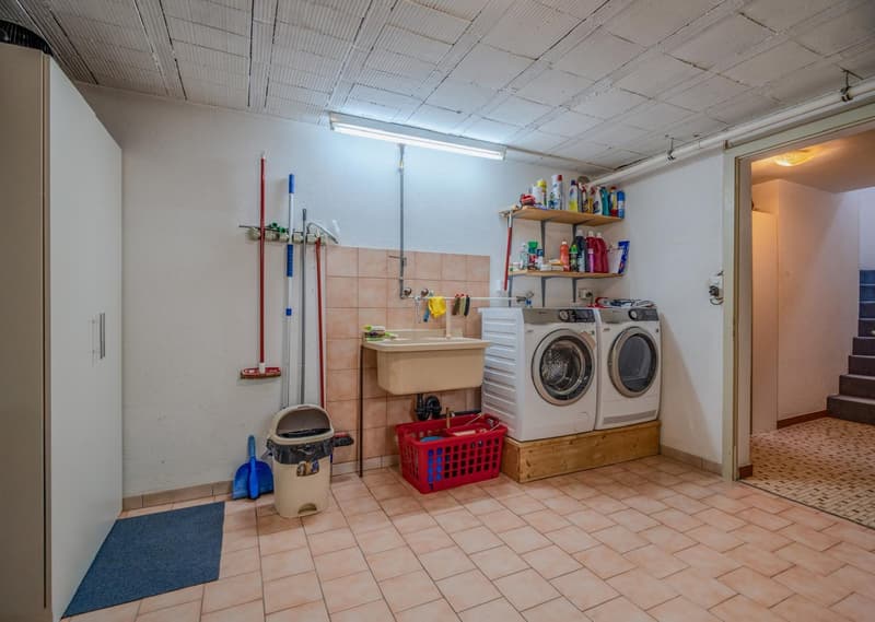 The image shows a utility room with a washing machine and dryer, a sink, shelves with cleaning supplies, and various cleaning tools and equipment. The room has a tiled floor and a drop ceiling.
