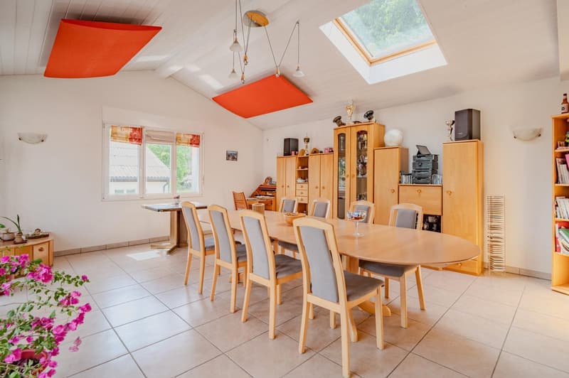 Spacious dining room with a large wooden dining table and chairs, wooden cabinets and shelves, and a skylight providing natural light. The room has a modern and bright feel with the orange accents on the ceiling.