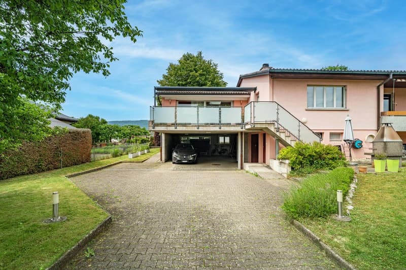 Two-story house with a garage under a balcony, pink exterior, brick driveway, front yard with bushes and plants, parked car in the garage
