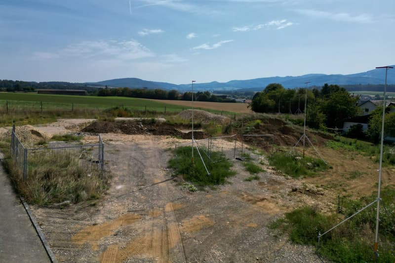 The image shows a construction site in a rural area. The site is surrounded by fencing and appears to be in the early stages of development, with exposed soil and construction equipment visible. The landscape in the background includes rolling hills, tree