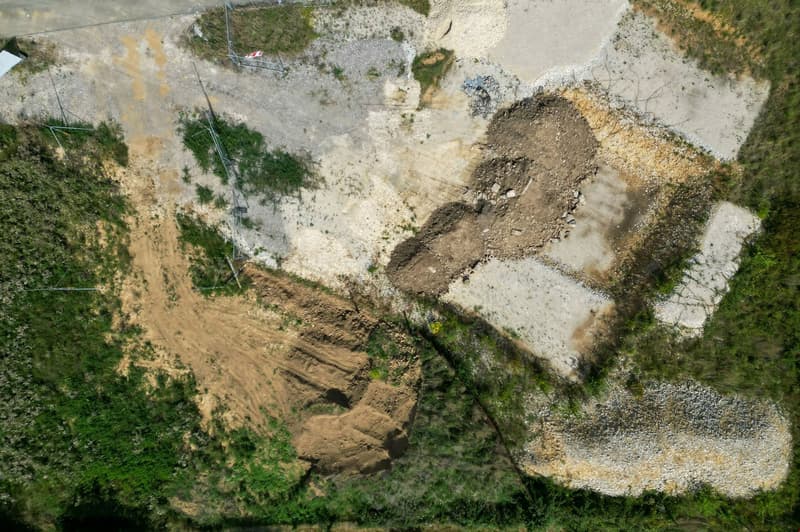 The image shows an aerial view of a rural or natural landscape. There is a dirt road or path winding through the area, with patches of vegetation and exposed soil or rock formations. The terrain appears to be uneven and rugged, with some areas of erosion