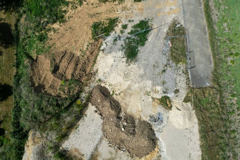 The image shows an aerial view of a rural area with a dirt road running through it. The landscape is a mix of grassy areas and bare, exposed soil. There are some trees and vegetation visible, but the overall scene appears to be a rural, undeveloped area.
