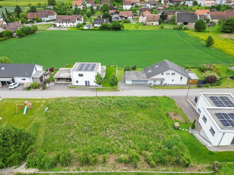 The image shows an aerial view of a residential area in a rural setting. The area consists of several houses with red-tiled roofs, surrounded by green fields and trees. There are two houses with attached garages and parking spaces visible. Additionally, s