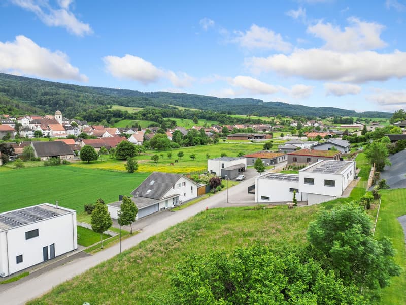 The image shows a picturesque rural village nestled in a lush, green valley surrounded by rolling hills and mountains. The village features a mix of traditional and modern buildings, with red-tiled roofs and white facades. The landscape is dotted with tre
