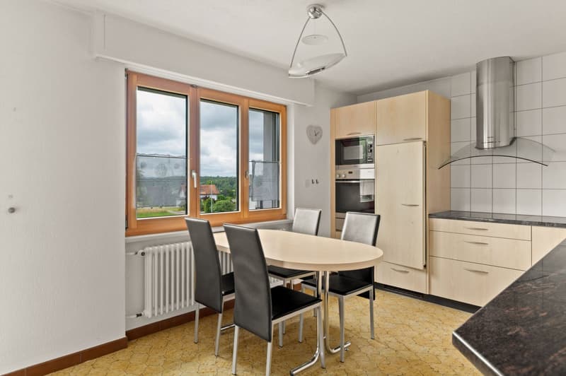 Spacious kitchen with large windows, wooden cabinets, stainless steel appliances including a dishwasher, and a dining table with chairs