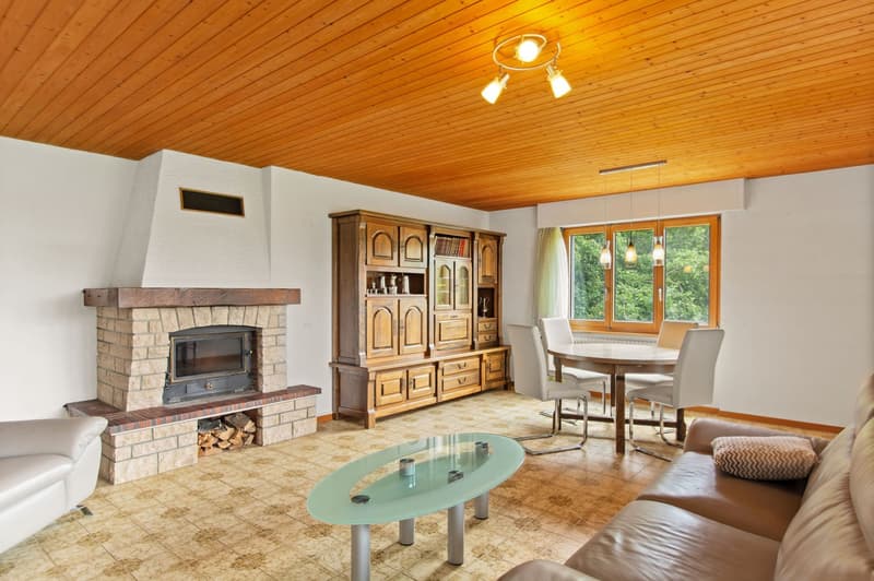 Spacious living room with a stone fireplace, wooden ceiling, large windows overlooking the outdoors, a wooden cabinet, a glass coffee table, and a dining area with a table and chairs.