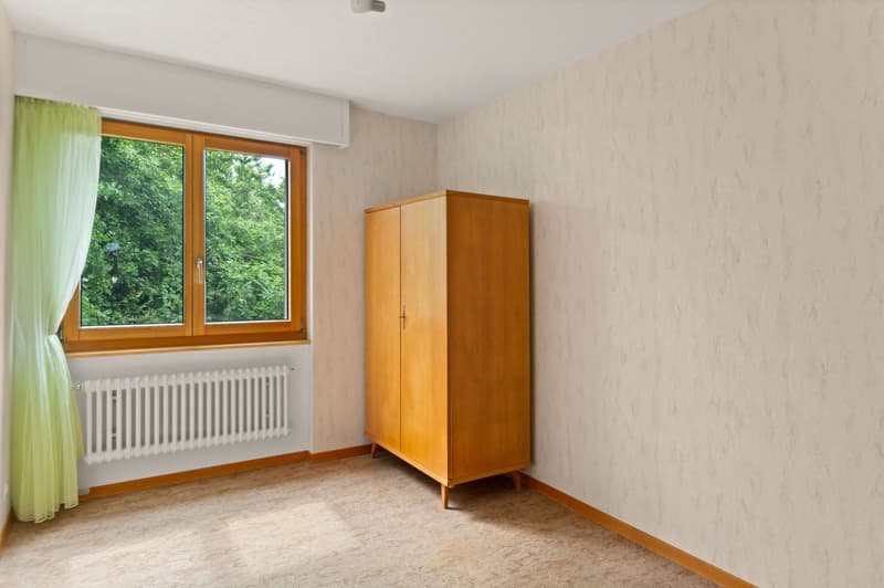 The image shows a bedroom with a large wooden wardrobe, a window overlooking greenery, and light-colored walls. The room has a carpeted floor and green curtains.
