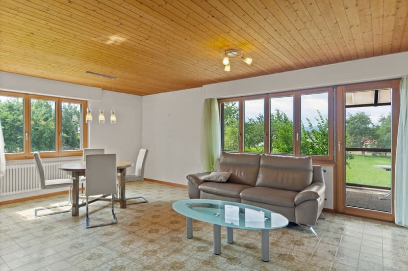 Spacious living room with large windows overlooking a garden, wooden ceiling, leather sofa, glass coffee table, and dining table with chairs.