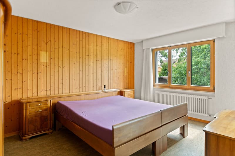 Spacious bedroom with wooden paneled walls, large windows overlooking greenery, a wooden bed frame with a purple bedspread, and a wooden nightstand.