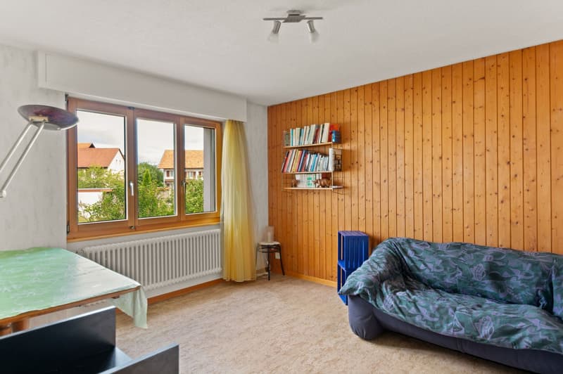 Spacious bedroom with wooden paneled walls, large windows providing a view, a bed with patterned bedding, a bookshelf, and a desk or table.