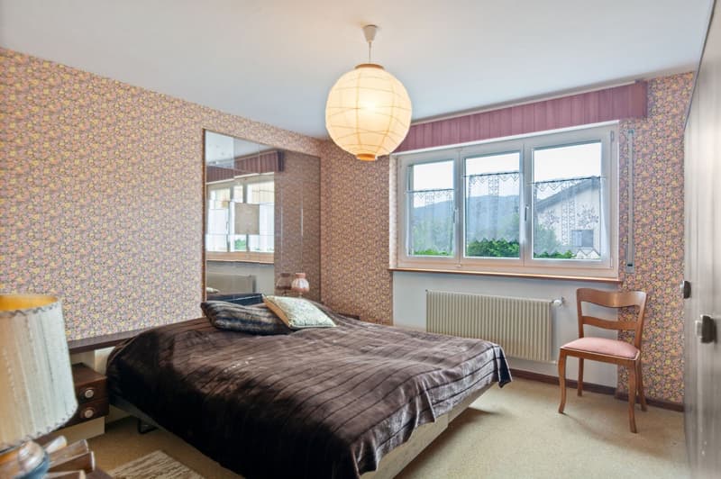 Spacious bedroom with floral wallpaper, large windows, a pendant light fixture, a double bed with a dark comforter, and a wooden chair.