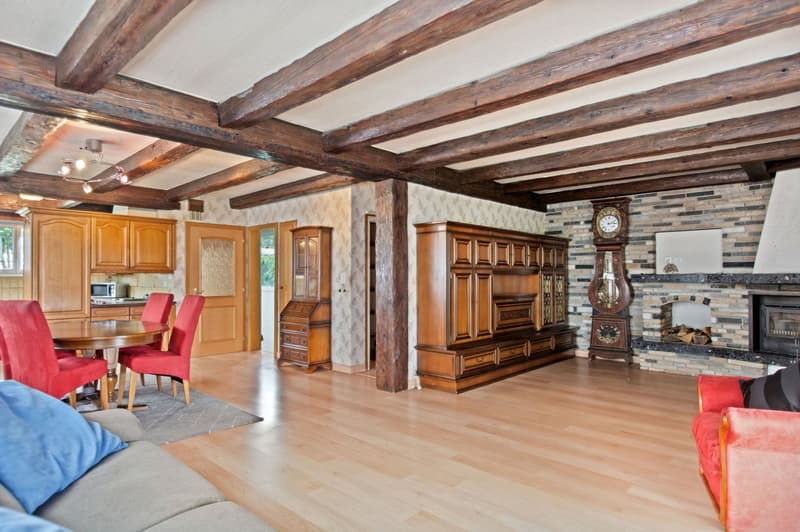 Large open-concept living space with exposed wooden beams on the ceiling, wooden cabinetry and furniture, a stone fireplace, and red upholstered chairs around a dining table