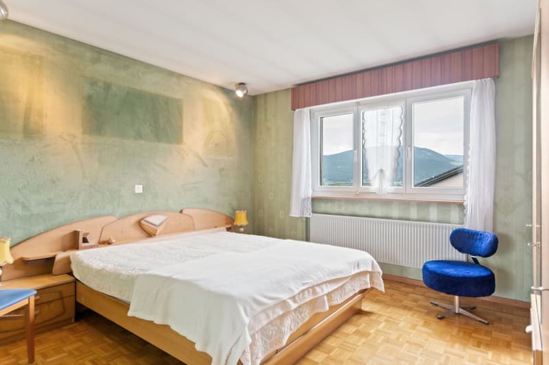 Spacious bedroom with large windows offering a scenic view, wooden floor, white bedding, wooden furniture including a bed frame and side tables, and a blue upholstered chair.