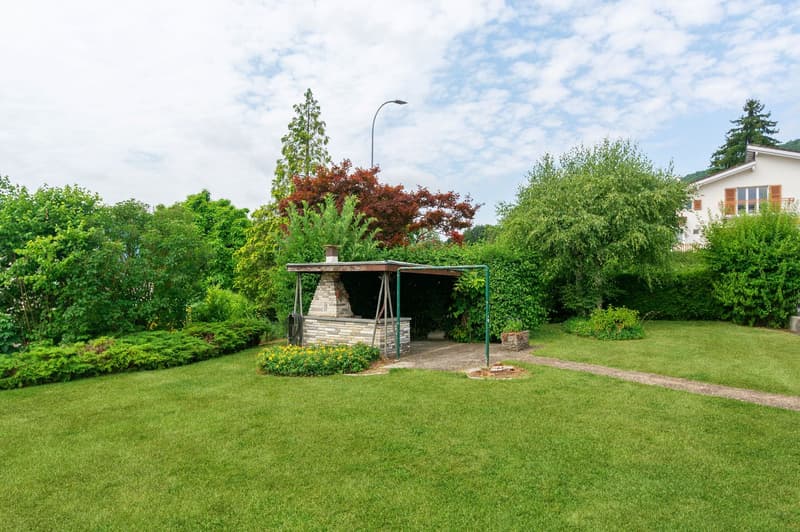 The image shows a well-maintained outdoor area with a lush green lawn, various trees and shrubs, and a stone gazebo structure with a roof. The surrounding area appears to be a residential neighborhood with other buildings visible in the background.
