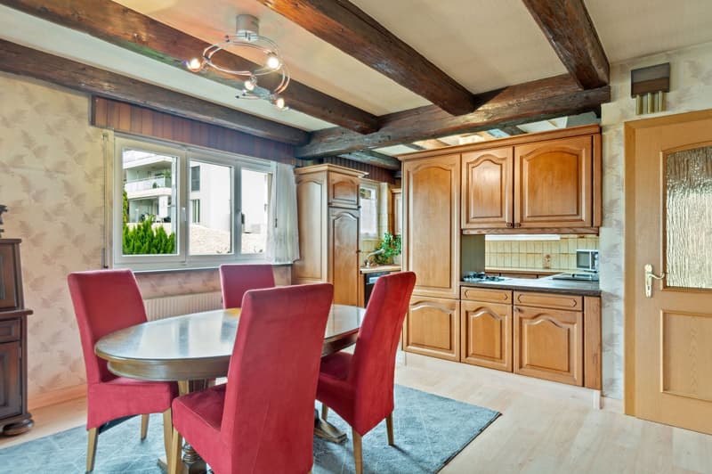 The image shows a combined kitchen and dining area with wooden beams on the ceiling, wooden cabinetry, a round dining table with red upholstered chairs, and a window overlooking the outdoors. The kitchen area features a built-in dishwasher.