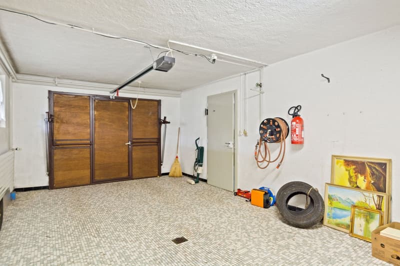 The image shows a garage space with wooden garage doors, a fire extinguisher, and various tools and equipment stored in the room. The floor is covered with a tiled mosaic pattern.