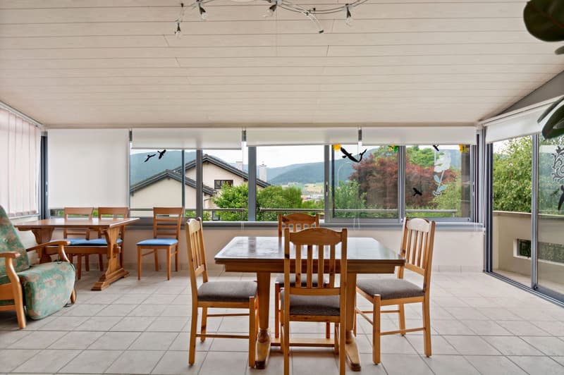 Spacious dining area with large windows offering a scenic view of the surrounding mountains and greenery. The room features a wooden dining table with chairs, as well as a couch and additional seating. The ceiling has a unique lighting fixture.